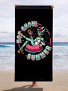 A beach towel with a gothic themed design featuring a character and the text 'Hot Ghoul Summer'. The towel is black with a graphic center, displayed at a beach setting.